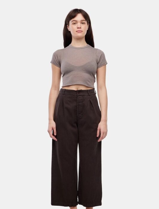 A woman stands facing forward wearing a short-sleeved, gray crop top, high-waisted dark wide-leg pants, and black flip-flops against a plain white background.