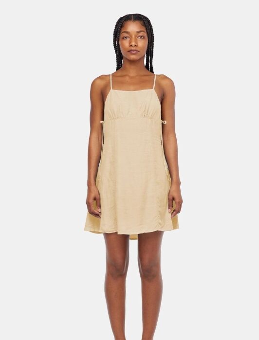 A woman stands facing forward wearing a sleeveless, light beige, knee-length dress with thin straps and black sandals against a plain white background.