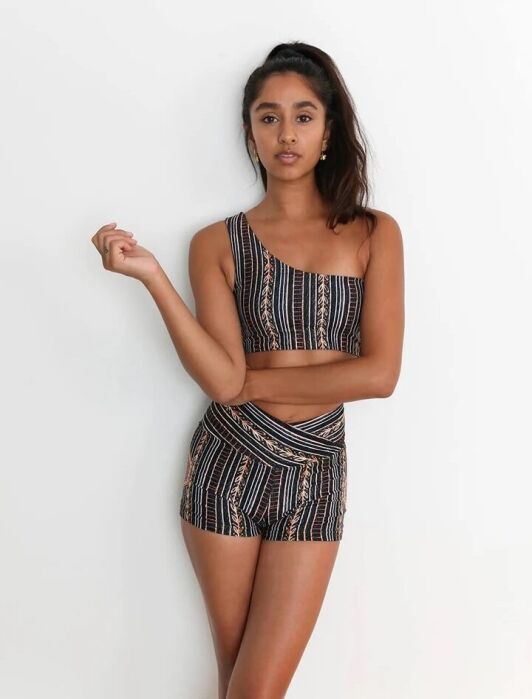 Woman standing against a white wall wearing a matching set of a one-shoulder crop top and high-waisted shorts with a black, orange, and white geometric pattern.