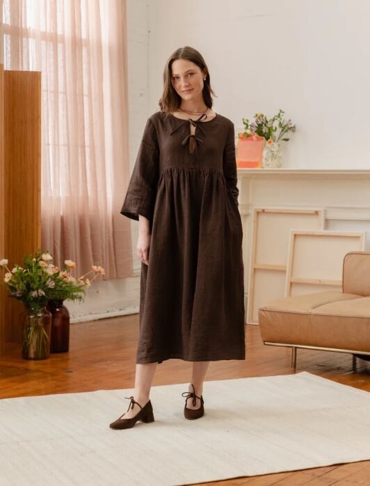 Woman stands indoors on a white rug, wearing a loose-fitting brown dress with a keyhole neckline and matching brown shoes. She is in a room with flowers, a couch, and large windows.