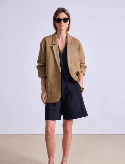 A woman wearing sunglasses, a tan blazer, black shirt, black shorts, and brown sandals stands against a plain background with one hand in her pocket.