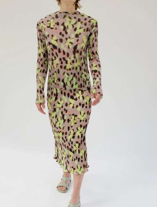 Person wearing a long-sleeved, midi-length dress with a green, pink, and black abstract pattern, and light green open-toe sandals, standing against a plain white background.