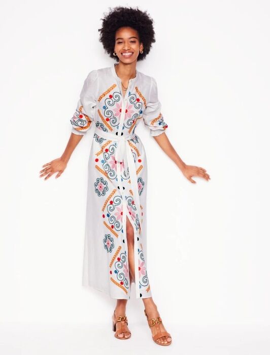 A woman stands against a white background wearing a long white shirt dress with colorful patterns, rolled-up sleeves, a waist tie, and tan sandals.