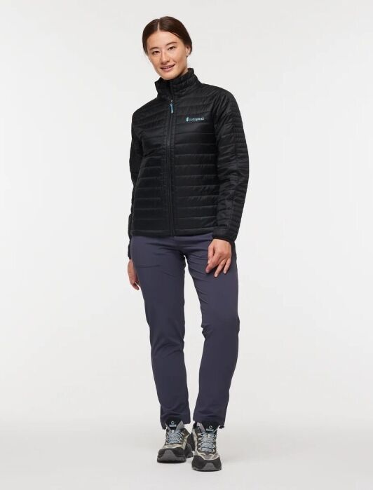 A person stands facing forward, wearing a black zip-up jacket, dark pants, and hiking shoes against a plain white background.