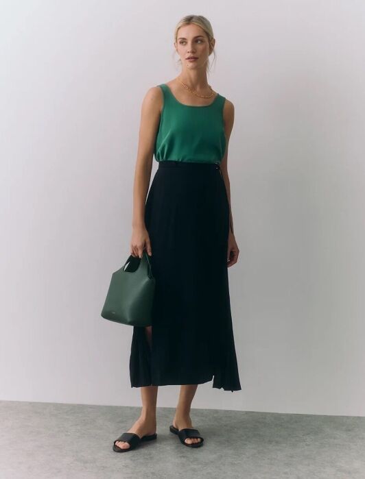A woman stands against a plain background wearing a green sleeveless top, black midi skirt, black sandals, and holding a small green handbag.
