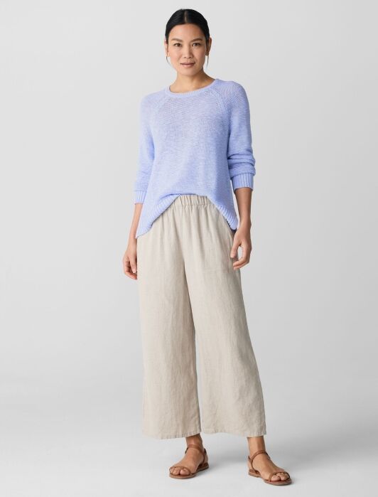A woman stands in a light blue sweater, beige wide-leg pants, and brown sandals against a plain light background.