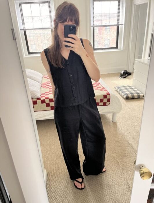 Person wearing a black sleeveless top and wide-leg pants takes a mirror selfie in a bedroom with a red and white checkered bedspread and a dog lying on the floor.