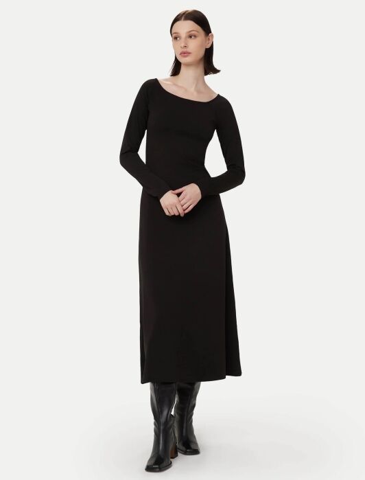 A woman stands wearing a long-sleeved, ankle-length black dress and black boots against a plain light background.
