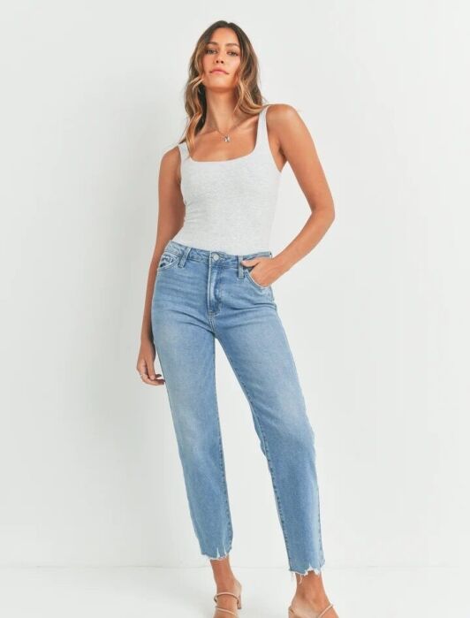 A woman stands against a white background wearing a light gray tank top, high-waisted blue jeans, and beige heeled sandals.
