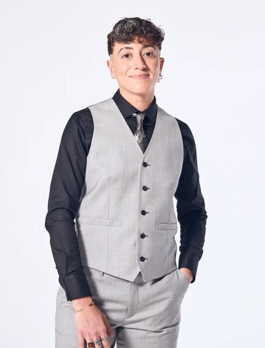 Person wearing a light gray vest and pants with a black shirt and patterned tie, standing against a plain white background with one hand in their pocket.
