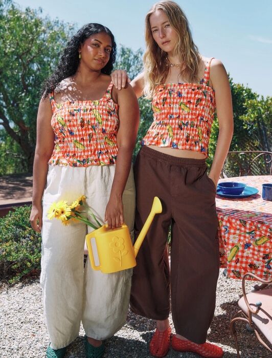 Two women stand outdoors wearing patterned orange tops; one holds a yellow watering can and sunflowers, both are dressed in loose pants. A table with a checkered tablecloth is in the background.