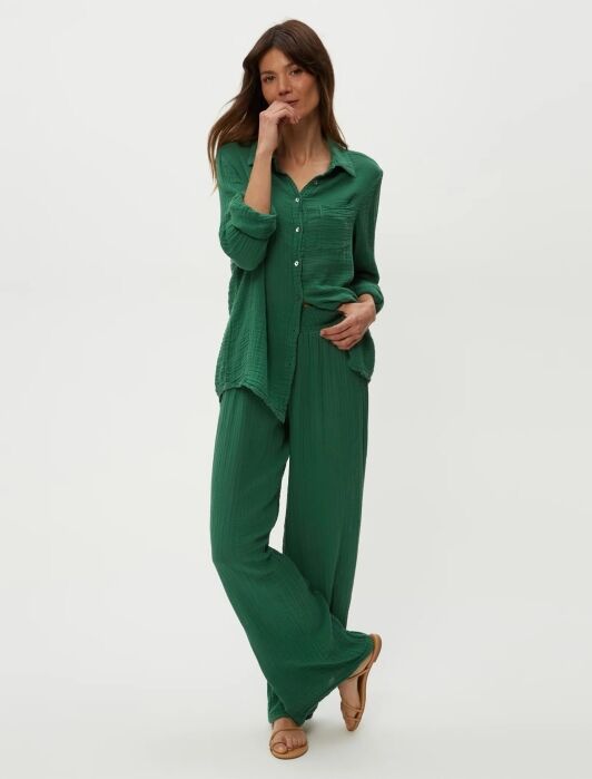 Woman standing against a plain light background, wearing a loose-fitting green button-up shirt and matching green wide-leg pants with tan sandals.
