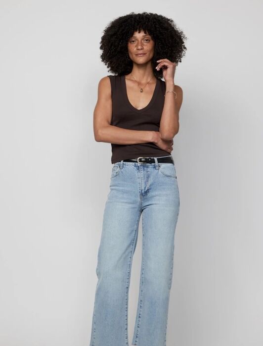 A person stands against a plain background, wearing a sleeveless black top, light blue jeans, and black shoes, with one hand touching their hair.