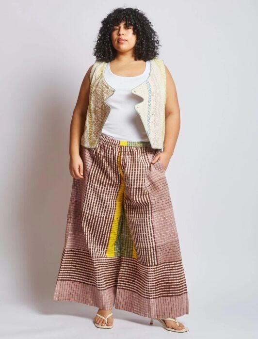 A woman stands against a plain background wearing a white tank top, a patterned vest, wide-legged checkered pants with yellow accents, and sandals.