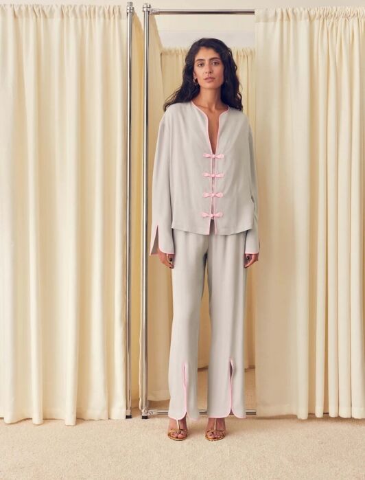 A woman stands in front of cream curtains, wearing a light gray pantsuit with pink trim and Mandarin-style buttons, and open-toe heels.
