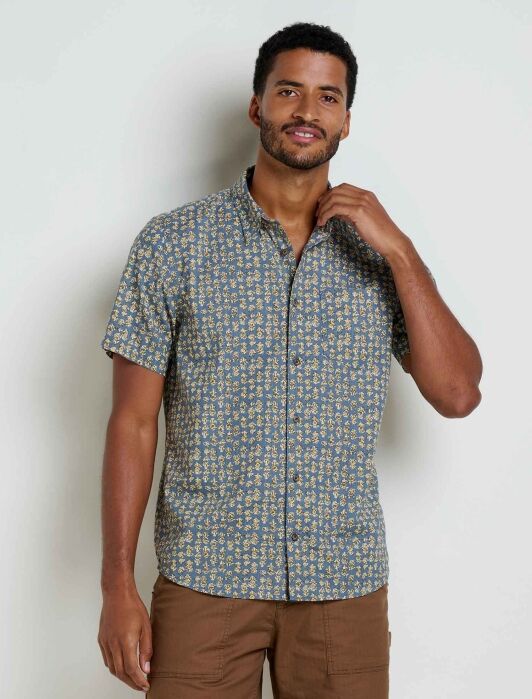 A man stands against a plain light-colored wall wearing a short-sleeve, button-up shirt with a small floral print and light brown pants.
