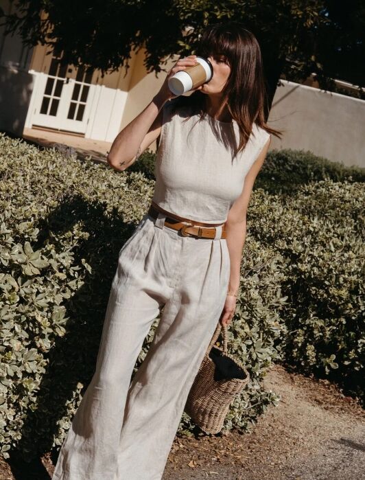 Woman stands outside on a sunny day, drinking from a takeaway cup. She wears a sleeveless beige jumpsuit, black sandals, and holds a woven bag.