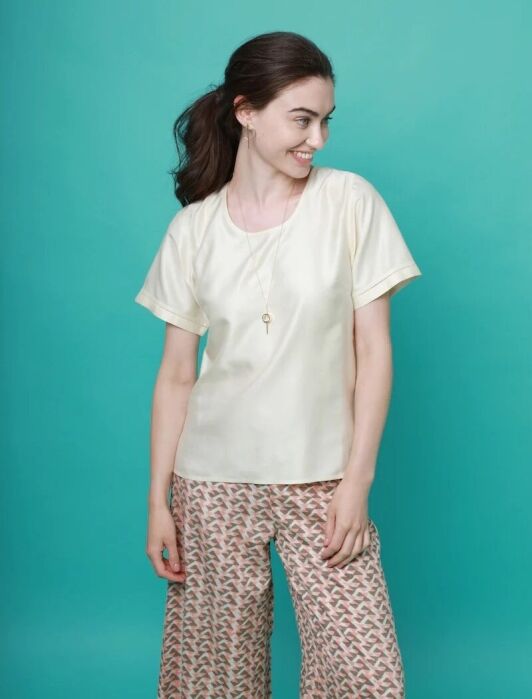 Woman standing in front of a teal background, wearing a light cream short-sleeve top, patterned wide-leg pants, and a necklace, looking to her left and smiling.