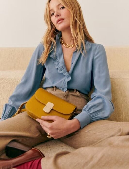 Woman sitting on a beige couch wears a blue blouse, tan trousers, and holds a mustard yellow handbag on her lap.