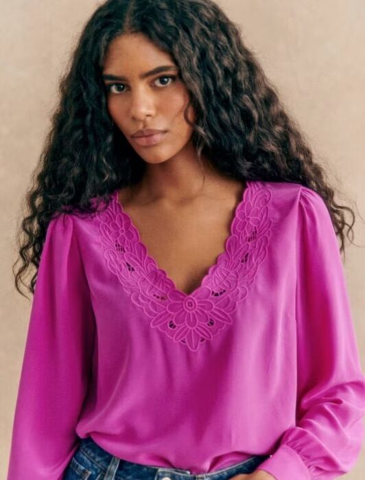Woman with long curly hair wearing a bright pink blouse with embroidered floral details at the V-neck, paired with blue jeans, standing against a neutral background.