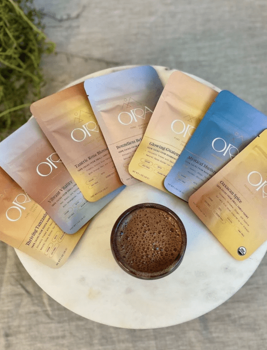 A round table displays a cup of frothy coffee surrounded by six colorful ORA coffee packets arranged in a semicircle.