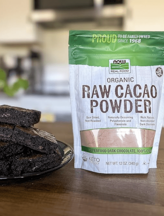 A plate of brownies sits beside a standing bag of NOW Real Food Organic Raw Cacao Powder on a kitchen counter.