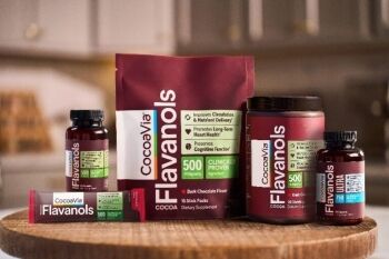 Various CocoaVia Flavanols supplement products, including bottles, a jar, a bag, and stick packs, are displayed on a wooden surface in a kitchen setting.