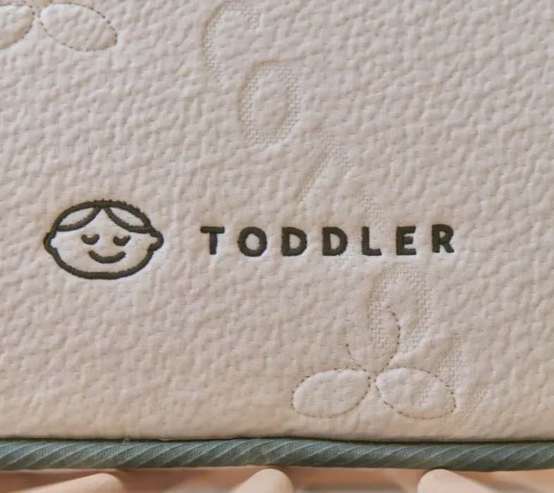 Close-up of a mattress label with a simple drawing of a child’s face and the word "TODDLER" stitched onto the fabric.