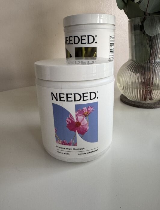 A white container labeled "NEEDED. Prenatal Multi Capsules" sits on a table, highlighting this premium prenatal vitamin; another similar container and a glass vase with green leaves appear in the background.