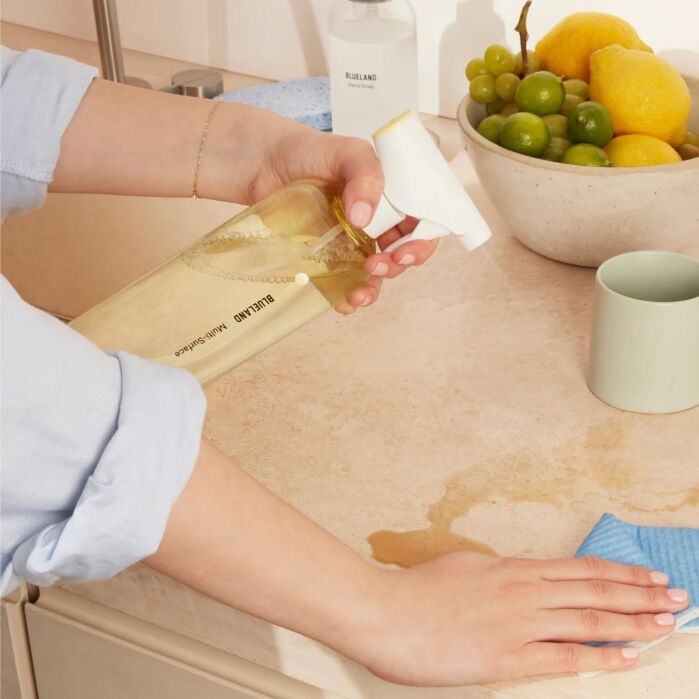 A person sprays a cleaning solution on a kitchen counter near spilled liquid, with a bowl of fruit and a green mug in the background.