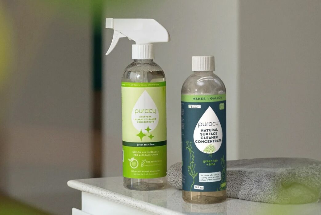 Two bottles of Puracy Natural Surface Cleaner Concentrate, one with a spray nozzle and one without, are placed on a counter next to a folded gray cloth.