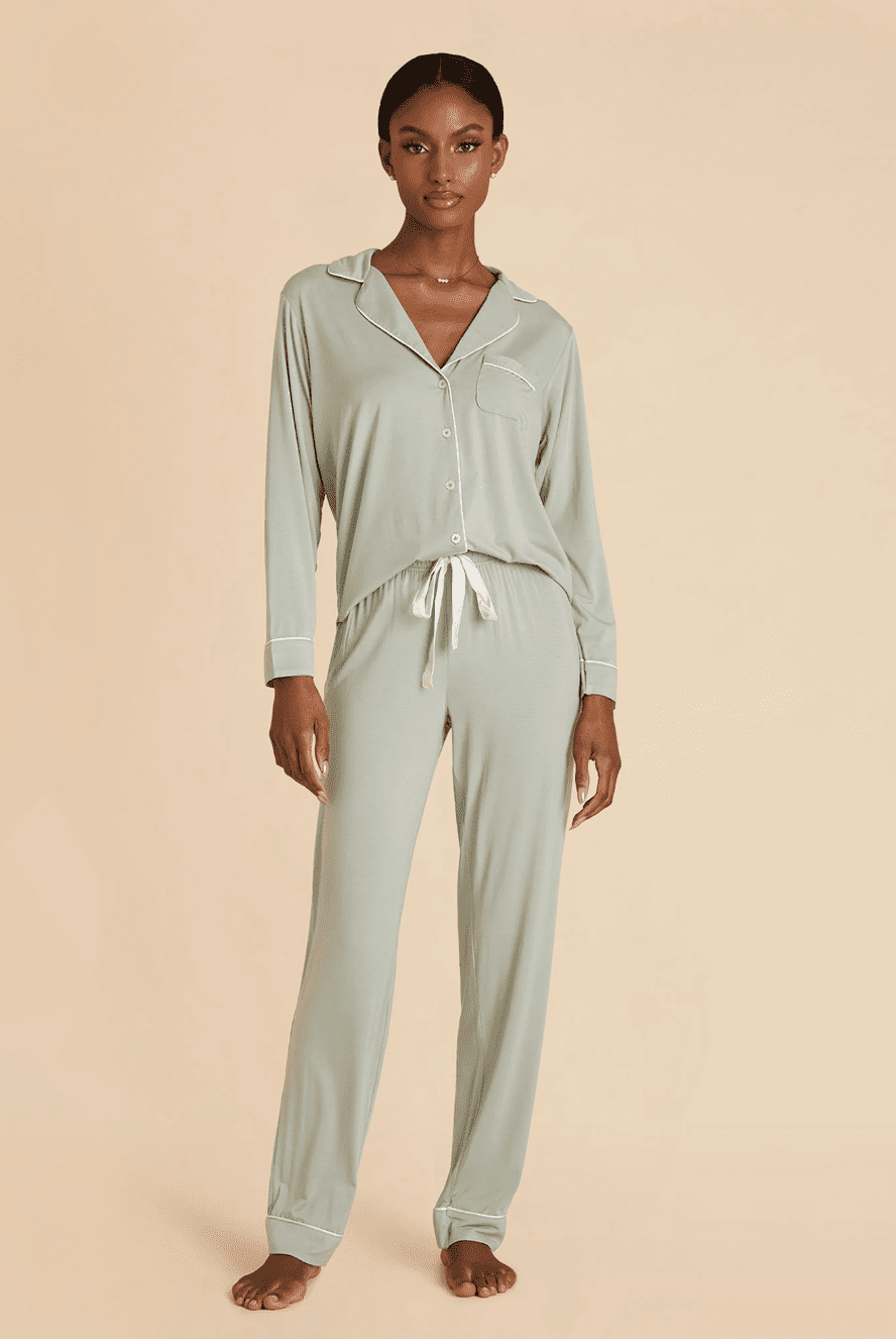 A woman stands barefoot wearing light sage green long-sleeve button-up pajamas with white trim and a drawstring waist, against a beige background.