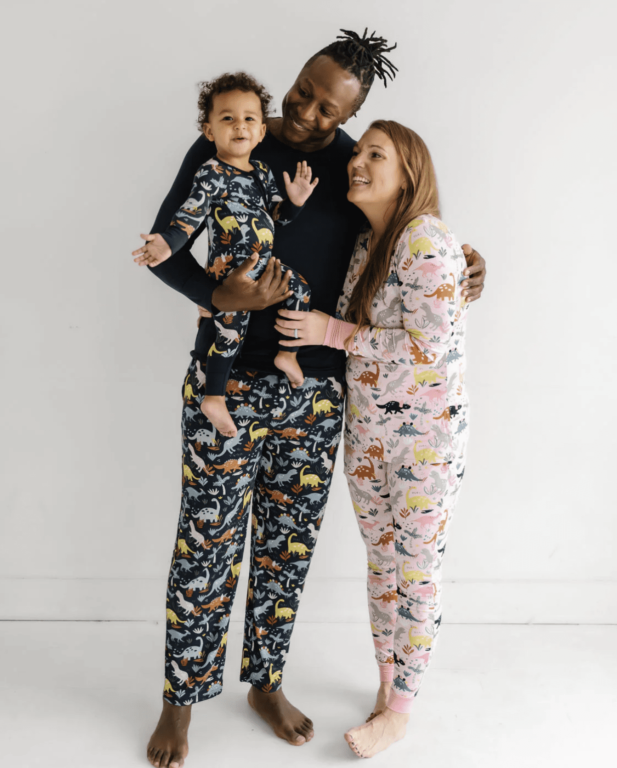 A family of three wearing dinosaur print pajamas stands together smiling against a plain white background.
