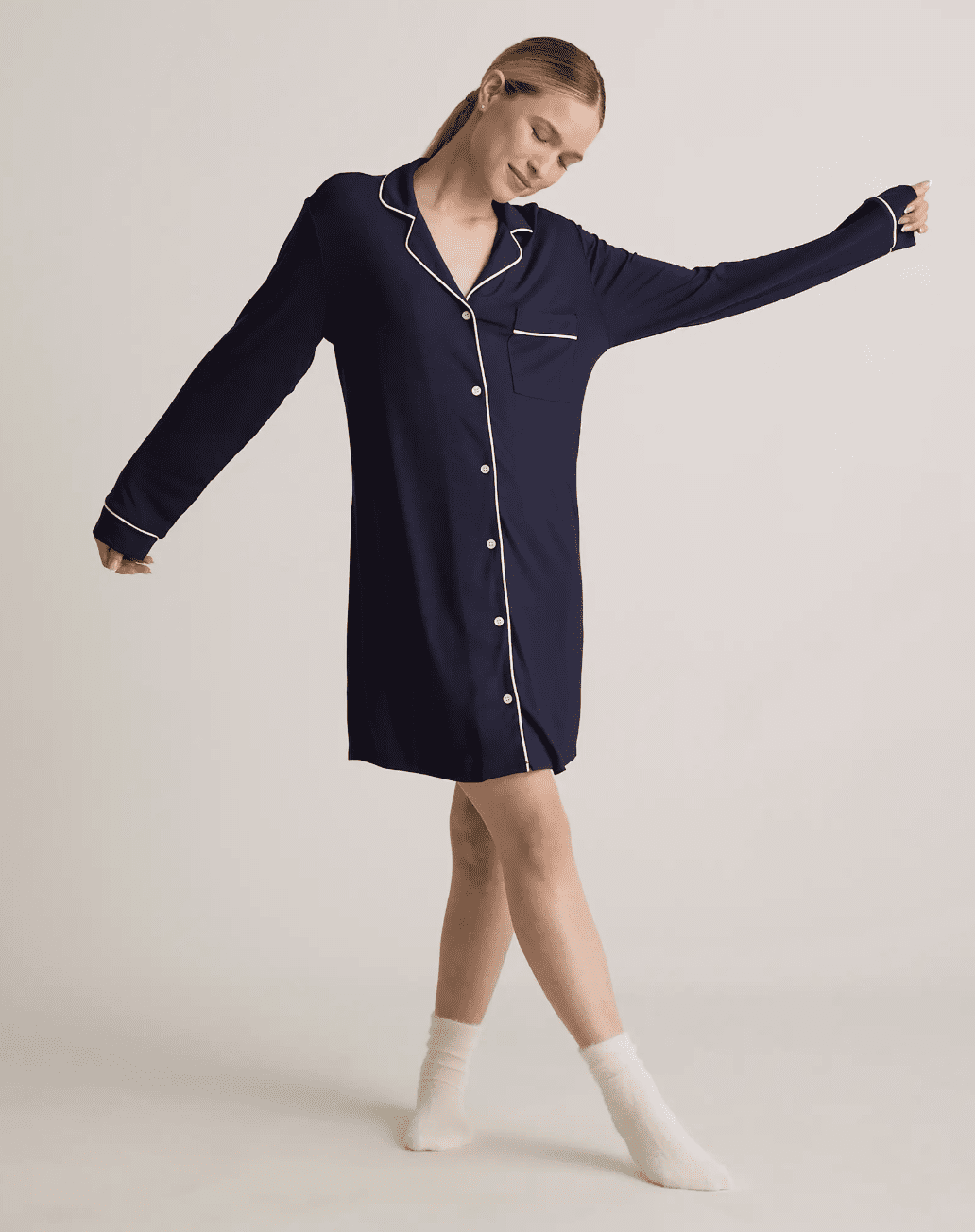 A woman wearing a long-sleeve, navy blue button-up nightshirt with white piping and white socks stands with arms extended against a plain background.
