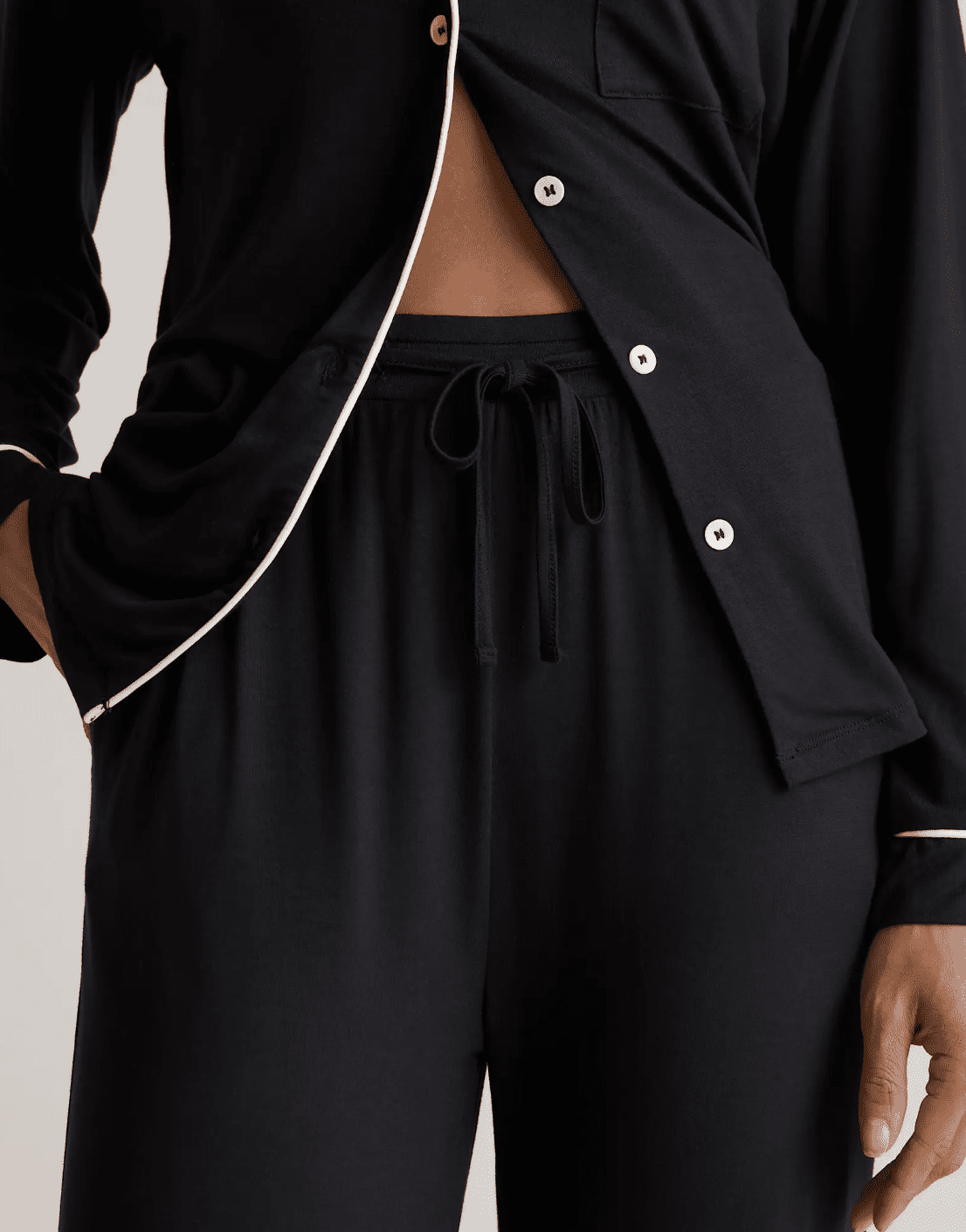 A person wearing black pajama pants and a matching black pajama shirt with white piping and buttons, one hand in their pocket.