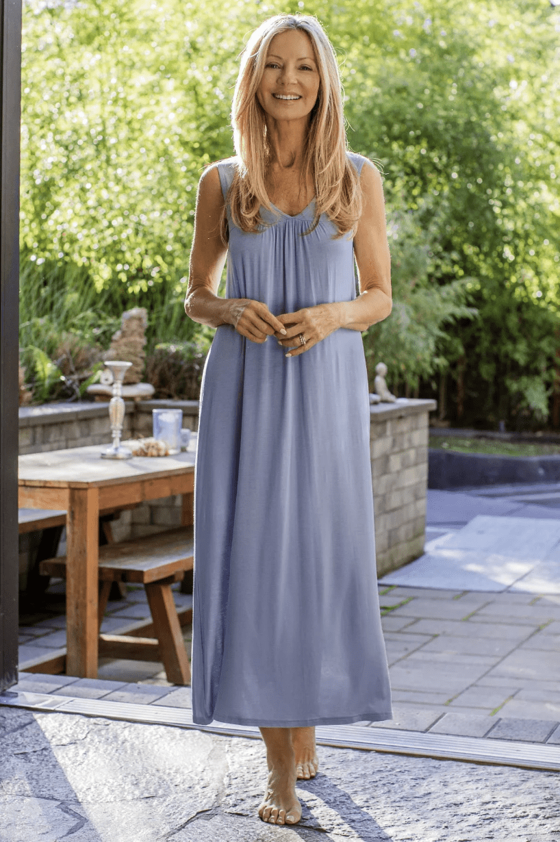 Woman with long blonde hair wearing a sleeveless light blue maxi dress stands barefoot on a patio with greenery and a wooden table in the background.