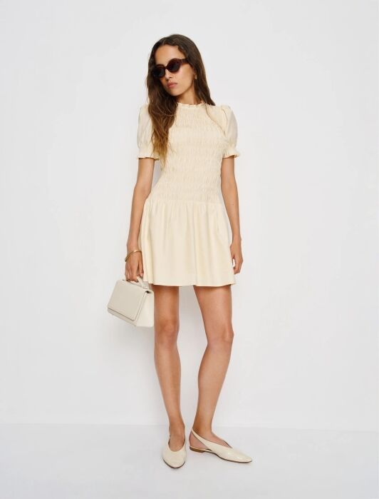 Woman wearing a cream-colored short dress with puff sleeves, cream flats, sunglasses, and holding a small cream handbag, channeling effortless French girl fashion against a plain white background.