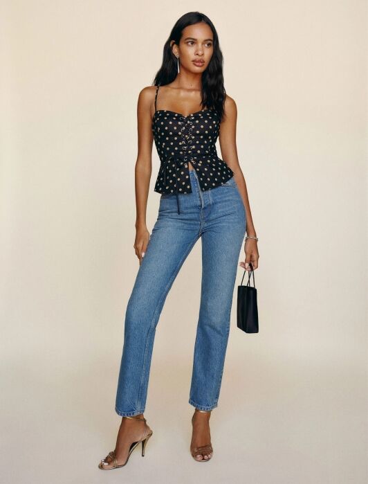 Woman standing against a beige background, channeling french girl fashion in a black floral top, blue jeans, heeled sandals, and holding a small black bag.