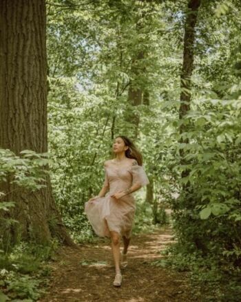 A woman in a light pink dress walks along a narrow, wooded path surrounded by dense green foliage and tall trees.