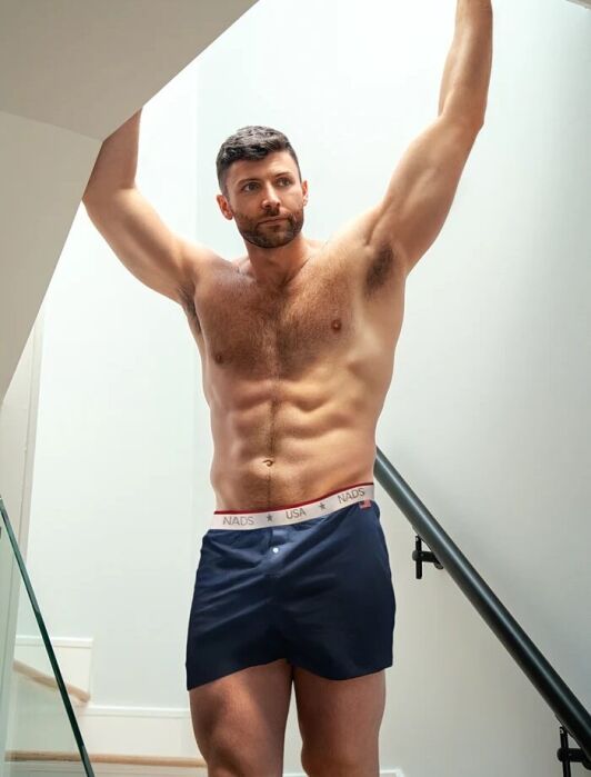 A shirtless man in navy blue boxers stands on a staircase, holding onto the ceiling with both arms raised.