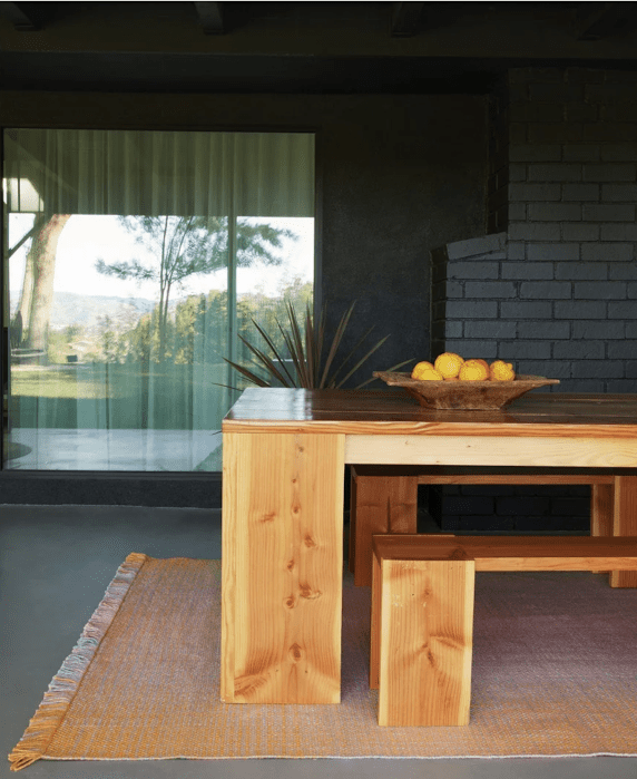 A wooden table with benches sits on a woven rug; a bowl of lemons is on the table. A large window and dark brick wall are in the background.