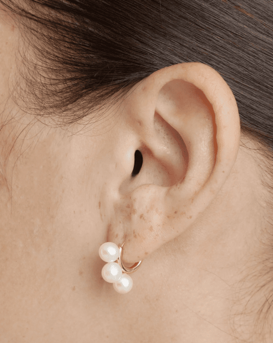 Close-up of a person’s ear wearing a gold hoop earring with three white pearls attached.