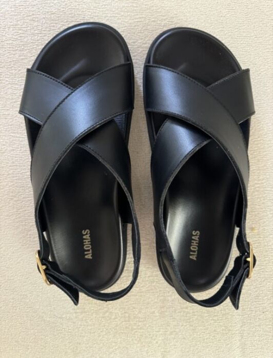 A pair of black sandals with crisscross straps and gold buckles, labeled "ALOHA" on the footbeds, placed on a light beige surface.