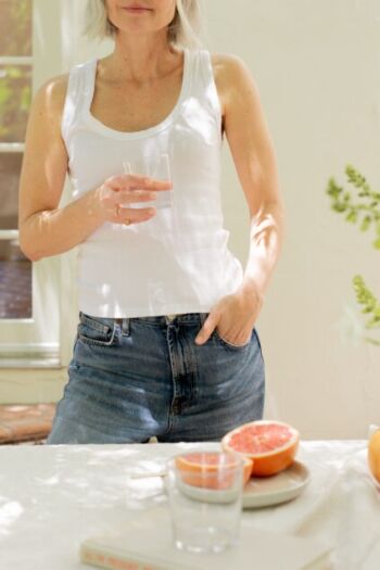 A person in a white tank top and jeans stands by a table holding a glass of water, with grapefruit halves and glasses on the table in sunlight.