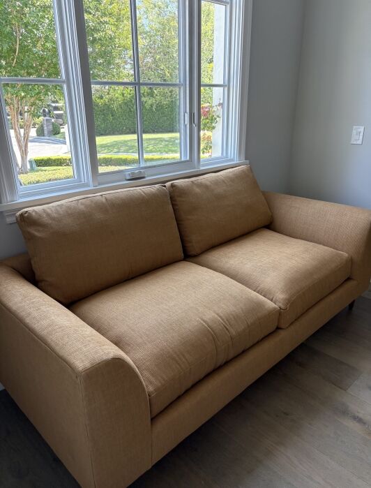 A tan fabric sofa with two seat cushions and two back cushions sits in front of a window with a view of greenery outside.