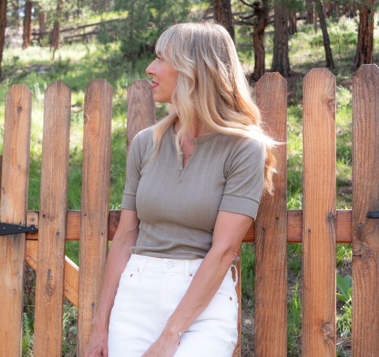 A woman with long blonde hair wearing a taupe short-sleeve top and white pants stands in front of a wooden fence, looking to the side. Trees and grass are in the background.