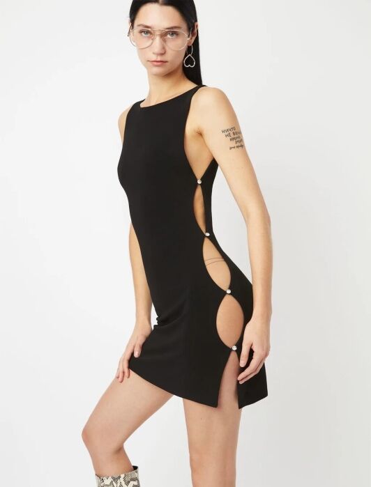 A woman wearing a sleeveless black mini dress with side cutouts fastened by metal rings, clear glasses, hoop earrings, and knee-high snakeskin boots embodies effortless French girl fashion against a plain white background.