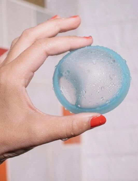 A hand with red painted nails holds a blue, round, textured object with water droplets on it against a tiled background.