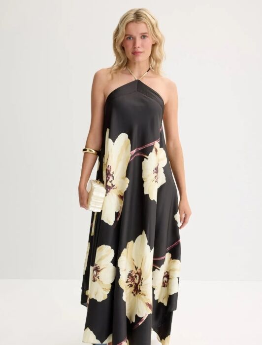 A woman stands against a plain background wearing a black halter-neck maxi dress with large yellow floral prints, gold sandals, a gold bracelet, and holding a small gold clutch.