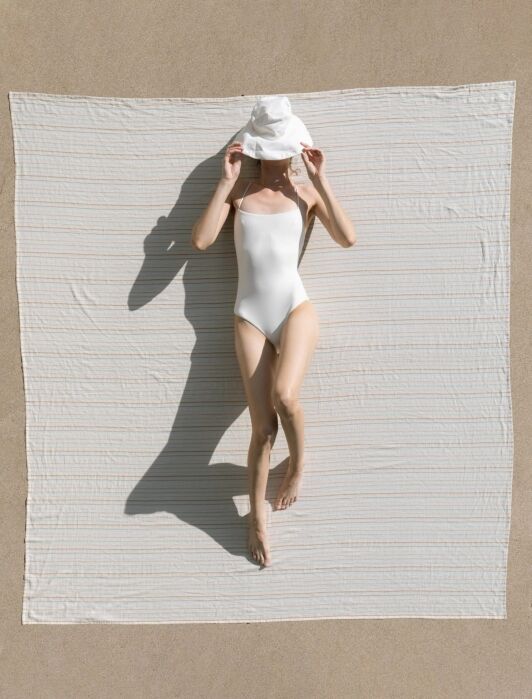A person in a white swimsuit and hat lies on a large white towel on sandy ground, casting a shadow beside them.